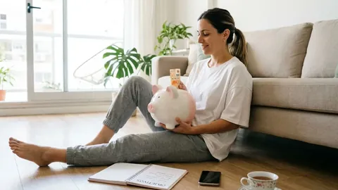 Foto de una mujer sentada en el piso del living que mete un billete en un chanchito blanco, con un cuaderno de objetivos