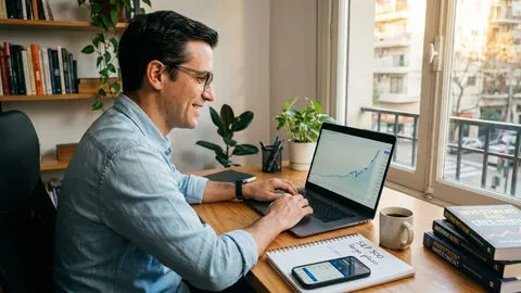 Foto de un hombre sonriente ante una laptop con un gráfico que sube, junto a una ventana, libros de inversión y un cuaderno